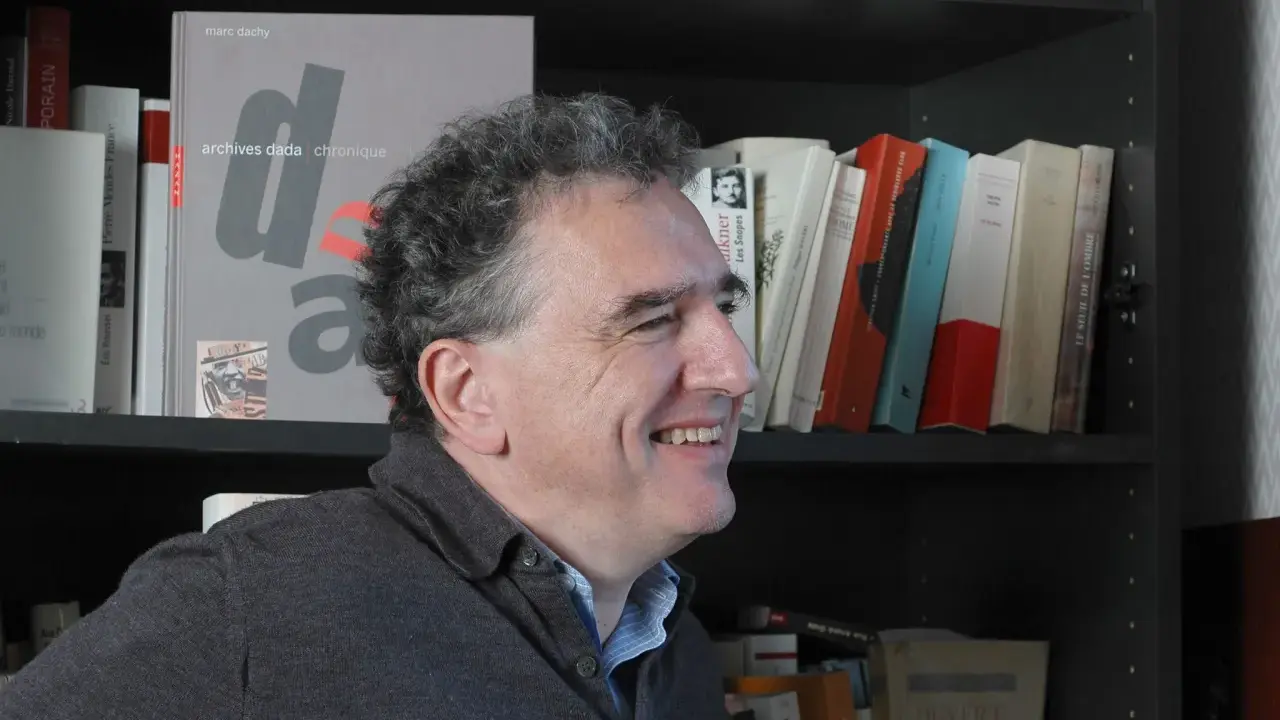 Un homme souriant devant une étagère remplie de livres, dont un intitulé "archives dada chronique". Une liste de magazine littéraire à portée de main.