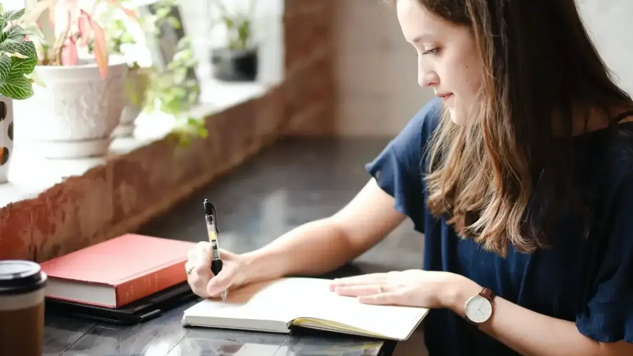 Jeune femme écrivant dans un carnet, entourée de plantes. Elle semble rédiger une biographie, concentrée sur son travail.
