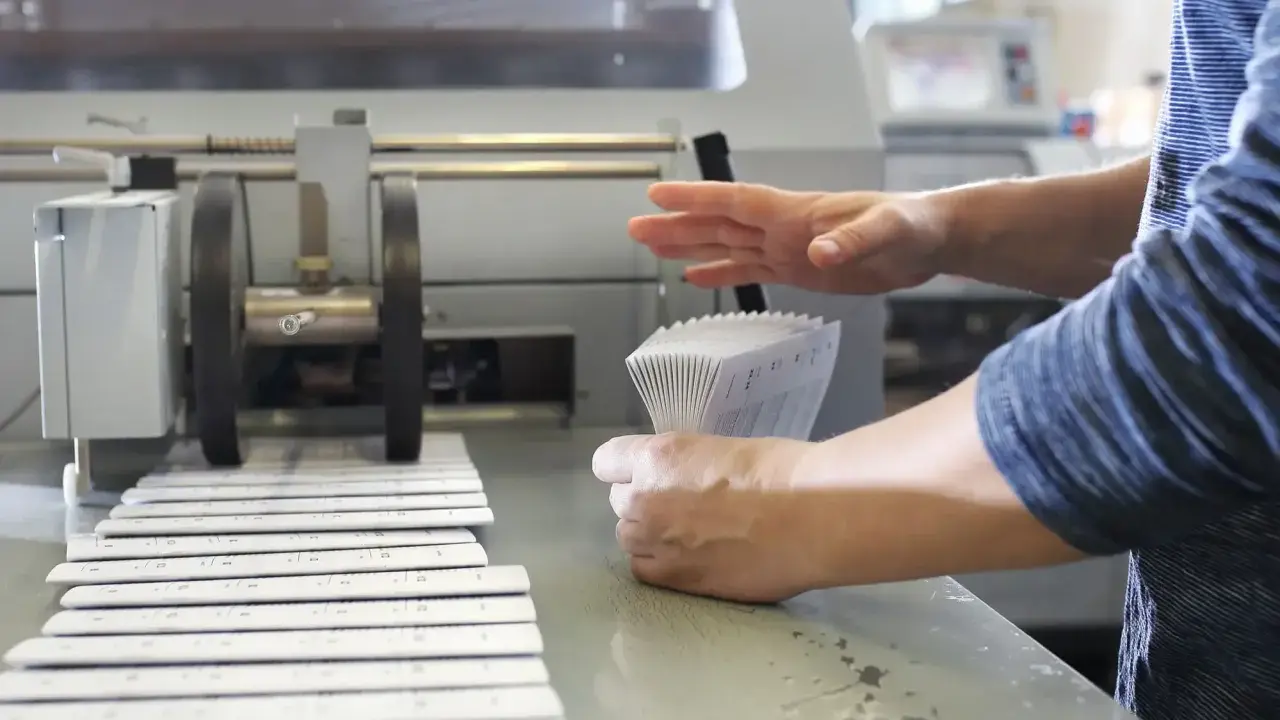 Mains manipulant une pile de documents imprimés pour le façonnage imprimerie, à côté d'une machine.