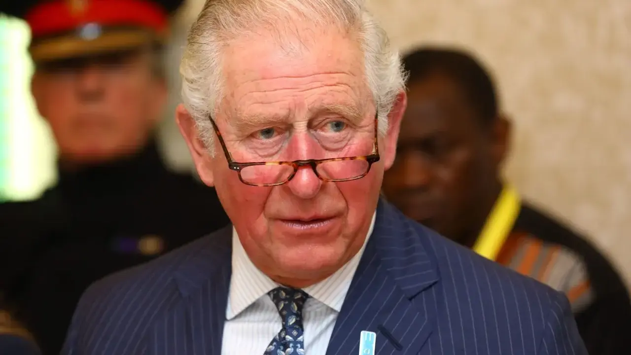 Un homme d'âge mûr, portant des lunettes et un costume rayé, semble réfléchir. Il pourrait être un membre du nom de famille royal imaginaire des Windsor.
