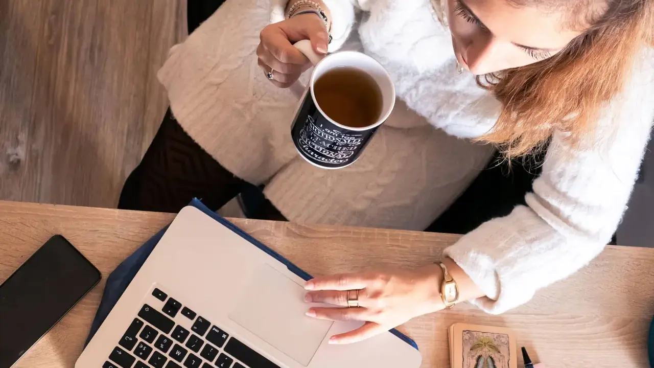 Une femme boit une tasse de thé en travaillant sur son ordinateur portable, entourée d'objets inspirants, peut-être pour ses **éditions ring manuscrit**.