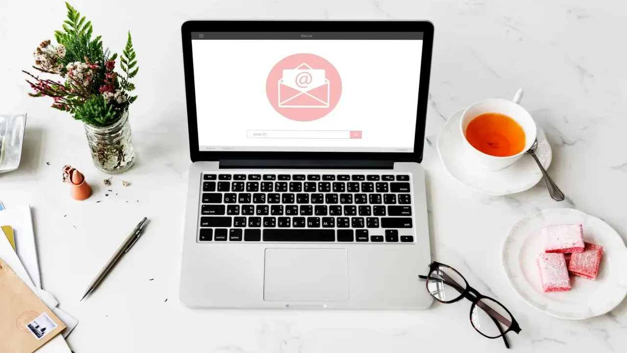 Un bureau avec un ordinateur affichant une enveloppe, une tasse de thé, des lunettes et des fleurs. Idéal pour un projet de pocket jeunesse manuscrit.