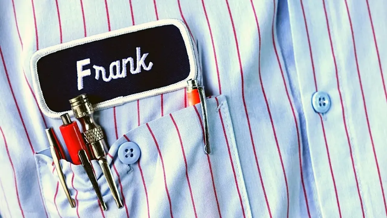 Chemise à rayures bleues et rouges avec une étiquette "Frank". Le prénom Frank est brodé en blanc. Dans la poche, des stylos et des outils.
