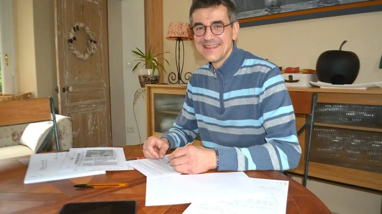 Un homme souriant, lunettes, pull rayé, travaille à son bureau. Il est en train de devenir écrivain public, prêt à rédiger vos textes.