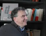 Un homme souriant devant une étagère remplie de livres, dont un intitulé "archives dada chronique". Une liste de magazine littéraire à portée de main.