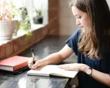 Jeune femme écrivant dans un carnet, entourée de plantes. Elle semble rédiger une biographie, concentrée sur son travail.