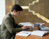 Un étudiant concentré sur son ordinateur portable, entouré de livres sur la rédaction de thèse. Il se prépare peut-être à la soutenance de thèse, se demandant combien de temps dure une soutenance de thèse.