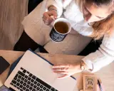 Une femme boit une tasse de thé en travaillant sur son ordinateur portable, entourée d'objets inspirants, peut-être pour ses **éditions ring manuscrit**.