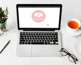 Un bureau avec un ordinateur affichant une enveloppe, une tasse de thé, des lunettes et des fleurs. Idéal pour un projet de pocket jeunesse manuscrit.