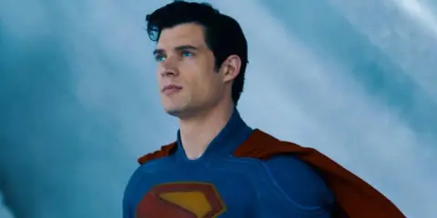 Superman, prénom de héros masculin, regarde au loin, son costume bleu et sa cape rouge flottant.