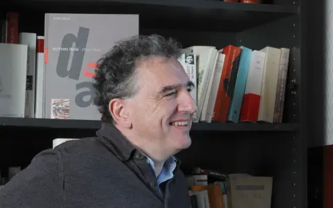 Un homme souriant devant une étagère remplie de livres, dont un intitulé "archives dada chronique". Une liste de magazine littéraire à portée de main.