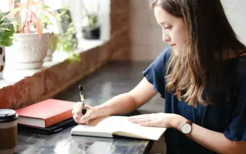 Jeune femme écrivant dans un carnet, entourée de plantes. Elle semble rédiger une biographie, concentrée sur son travail.