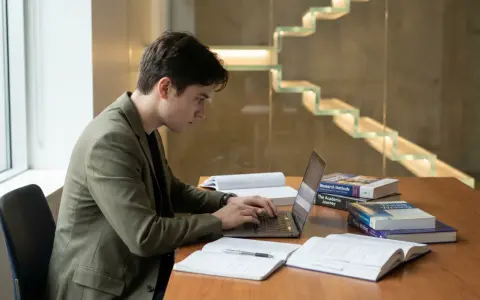 Un étudiant concentré sur son ordinateur portable, entouré de livres sur la rédaction de thèse. Il se prépare peut-être à la soutenance de thèse, se demandant combien de temps dure une soutenance de thèse.