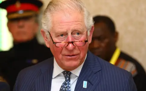 Un homme d'âge mûr, portant des lunettes et un costume rayé, semble réfléchir. Il pourrait être un membre du nom de famille royal imaginaire des Windsor.
