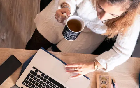 Une femme boit une tasse de thé en travaillant sur son ordinateur portable, entourée d'objets inspirants, peut-être pour ses **éditions ring manuscrit**.