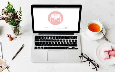 Un bureau avec un ordinateur affichant une enveloppe, une tasse de thé, des lunettes et des fleurs. Idéal pour un projet de pocket jeunesse manuscrit.