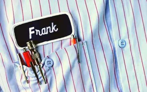 Chemise à rayures bleues et rouges avec une étiquette "Frank". Le prénom Frank est brodé en blanc. Dans la poche, des stylos et des outils.