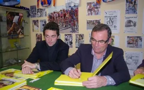 Deux hommes signent des livres jaunes sur le cyclisme, avec des affiches de la Martinière Jeunesse et des manuscrits en arrière-plan.