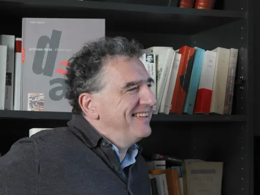 Un homme souriant devant une étagère remplie de livres, dont un intitulé "archives dada chronique". Une liste de magazine littéraire à portée de main.