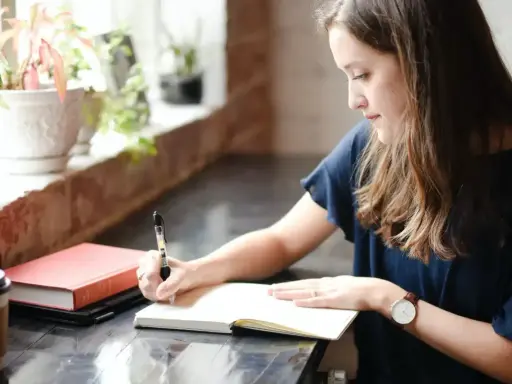 Jeune femme écrivant dans un carnet, entourée de plantes. Elle semble rédiger une biographie, concentrée sur son travail.