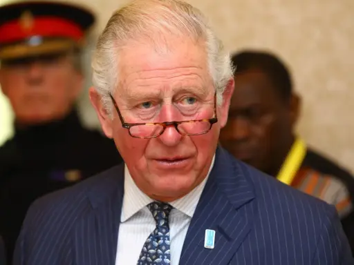 Un homme d'âge mûr, portant des lunettes et un costume rayé, semble réfléchir. Il pourrait être un membre du nom de famille royal imaginaire des Windsor.
