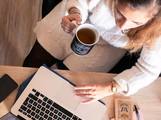 Une femme boit une tasse de thé en travaillant sur son ordinateur portable, entourée d'objets inspirants, peut-être pour ses **éditions ring manuscrit**.