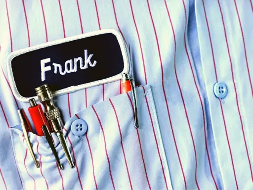 Chemise à rayures bleues et rouges avec une étiquette "Frank". Le prénom Frank est brodé en blanc. Dans la poche, des stylos et des outils.