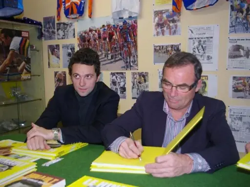 Deux hommes signent des livres jaunes sur le cyclisme, avec des affiches de la Martinière Jeunesse et des manuscrits en arrière-plan.