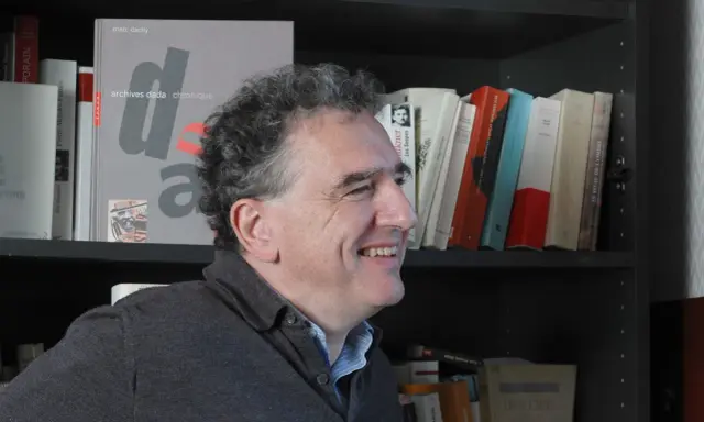 Un homme souriant devant une étagère remplie de livres, dont un intitulé "archives dada chronique". Une liste de magazine littéraire à portée de main.
