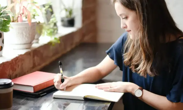 Jeune femme écrivant dans un carnet, entourée de plantes. Elle semble rédiger une biographie, concentrée sur son travail.