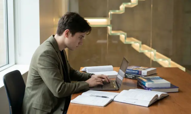 Un étudiant concentré sur son ordinateur portable, entouré de livres sur la rédaction de thèse. Il se prépare peut-être à la soutenance de thèse, se demandant combien de temps dure une soutenance de thèse.