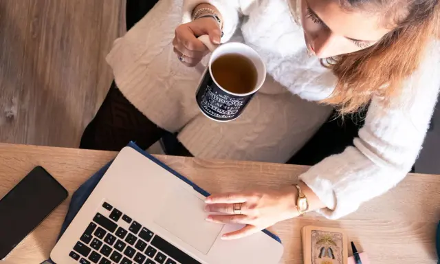 Une femme boit une tasse de thé en travaillant sur son ordinateur portable, entourée d'objets inspirants, peut-être pour ses **éditions ring manuscrit**.