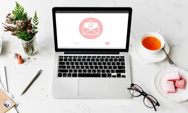 Un bureau avec un ordinateur affichant une enveloppe, une tasse de thé, des lunettes et des fleurs. Idéal pour un projet de pocket jeunesse manuscrit.