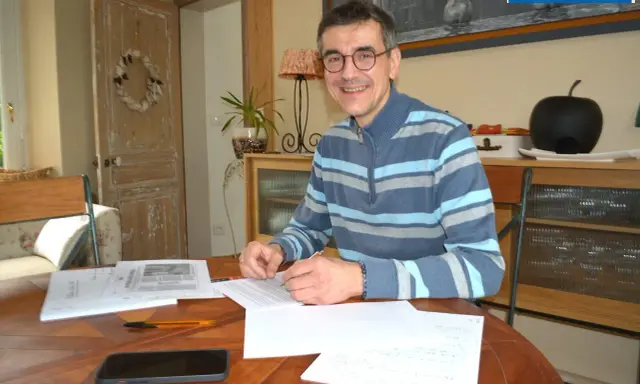 Un homme souriant, lunettes, pull rayé, travaille à son bureau. Il est en train de devenir écrivain public, prêt à rédiger vos textes.