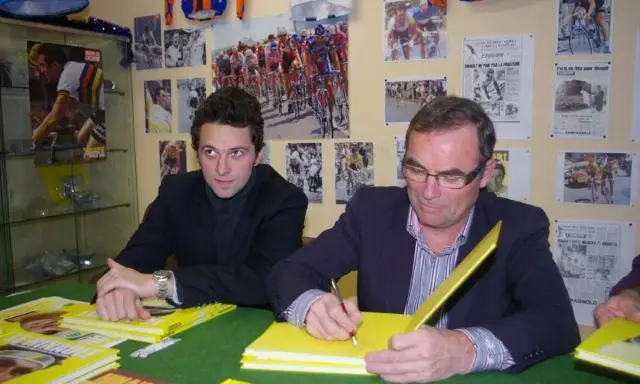 Deux hommes signent des livres jaunes sur le cyclisme, avec des affiches de la Martinière Jeunesse et des manuscrits en arrière-plan.