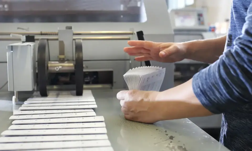 Mains manipulant une pile de documents imprimés pour le façonnage imprimerie, à côté d'une machine.