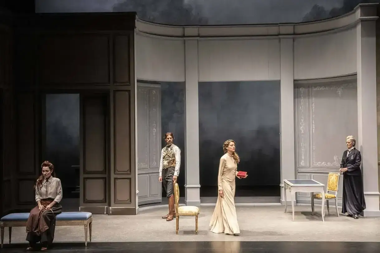 Sc&egrave;ne de th&eacute;&acirc;tre : quatre personnages dans un d&eacute;cor sobre. Une femme assise, un homme debout, une femme en robe longue tenant un livre, et une femme plus &acirc;g&eacute;e. Une &eacute;vocation des 