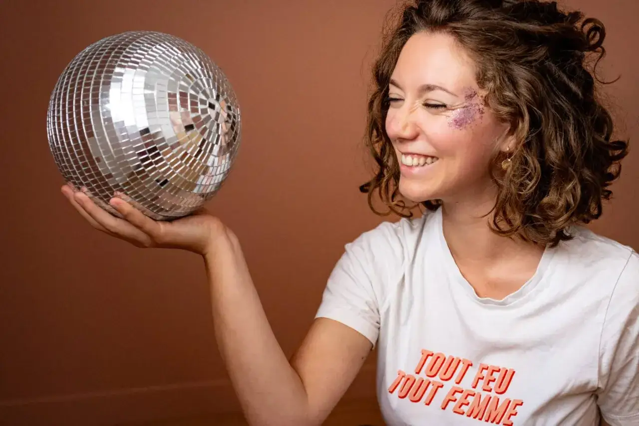 Une femme souriante, maquill&eacute;e de paillettes, tient une boule &agrave; facettes. Son t-shirt affiche 