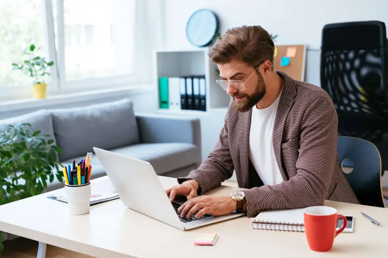 Homme concentr&eacute; sur son ordinateur portable, pr&ecirc;t &agrave; gagner de l'argent en &eacute;crivant. Bureau moderne avec tasse rouge et stylos color&eacute;s.